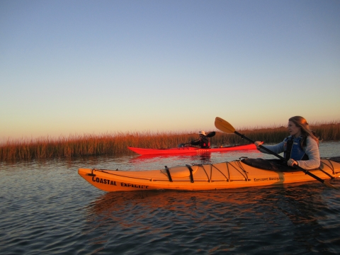 A woman and man are kayaking in the salt marsh estuary. The late afternoon sun casts a bright orange tint on the kayaks and the marsh grass behind the kayakers.