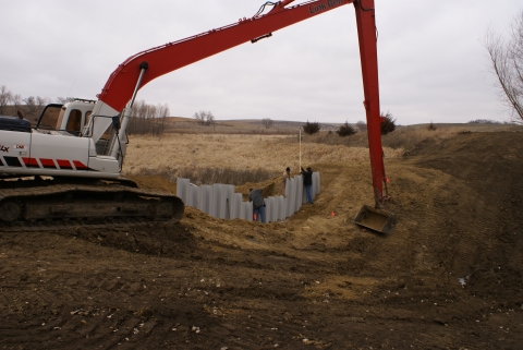 An excavator pushes in metal plates to form a wall on the outlet side of the wetland to help backup water in restore the wetland