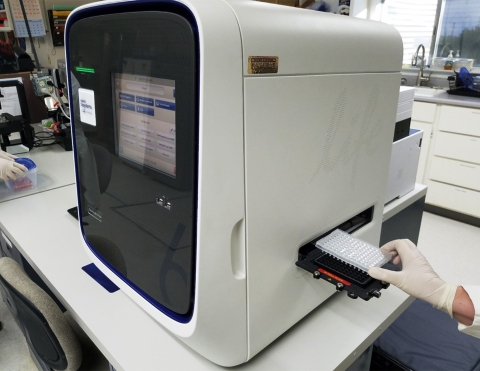 A large white qPCR machine has a visible screen on the front and a tray with multiple holes is being loaded into the side of the machine.  