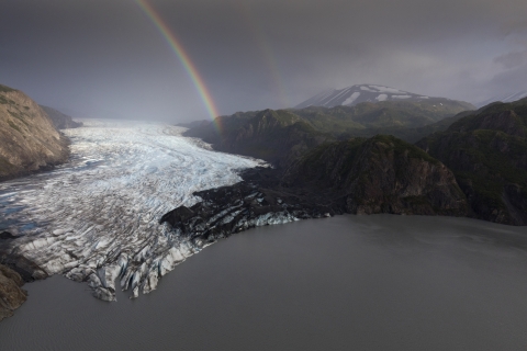 A rainbow arcs over a glacier into a green mountain range with a lake.
