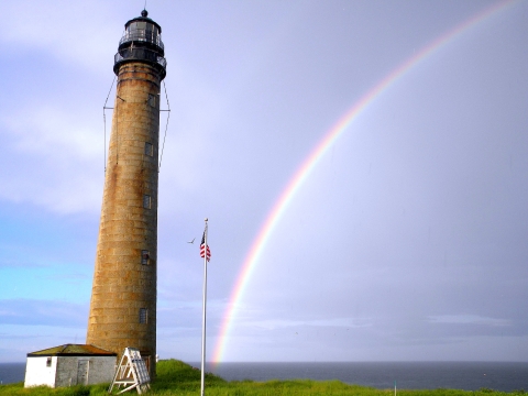 A tall, slender stone lighthouse on the coast with a rainbow over the ocean water