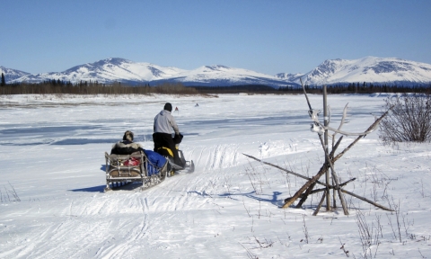 A man drives a snow mobile pulling a sled with a passenger in it across a snow-covered trail along a frozen river