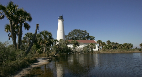 A  white lighthouse surrounded by palm trees in a spit of land near water