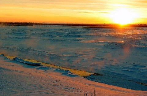 A snowy field bisected by a frigid river with the bright yellow sun shining just at the horizon
