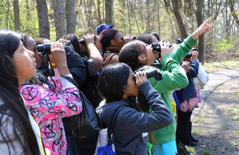 A dozen or so students on a wooded trail peer through binoculars and point to something unseen in the distance 