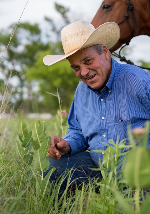 Bill Sproul, a rancher wearing a button-up blue shirt and ivory-colored cowboy hat, crouches in grass with his horse visible in the background