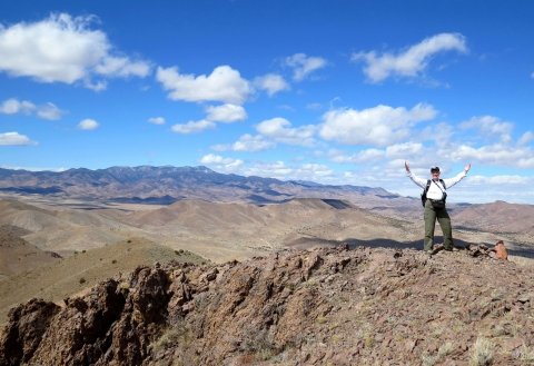 A man raises his arms in triumph atop a rocky mountain.