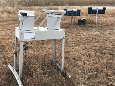 A solar-powered NDAP station, consisting of a metal frame with two bucket-shaped receptacles, collects precipitation data weekly throughout the year.