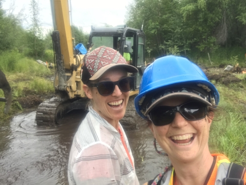 Two women in sunglasses and ballcaps stand in front of a piece of heavy equipment in water and grin for the camera.