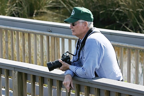 A gentleman on a walkway at Sabine NWR with a 35 millimeter camera in hand looking for his next subject to photograph