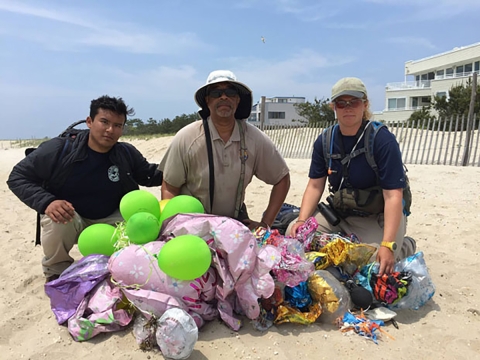 Three people, including one in Fish and Wildlife Service uniform, kneel on a beach alongside a pile of deflated balloons.