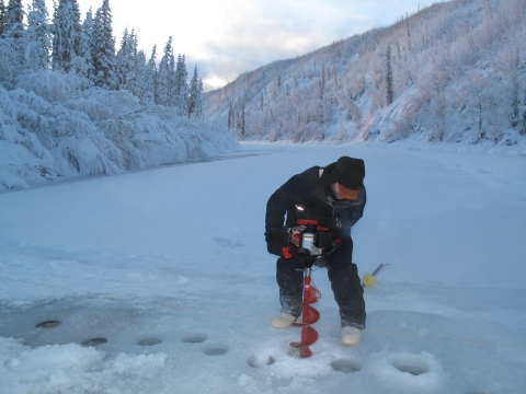 A hydrologist using power auger on ice covered water way during a streame gage project.