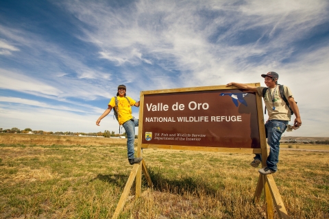 Two smiling young adults hang off a sign announcing Valle de Oro National Wildlife Refuge.