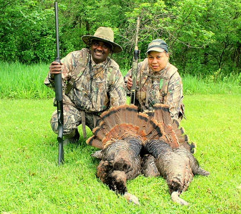 Wayne Hubbard and another hunter kneeling next to two turkey they hunted while wearing their fatigue and holding their rifles. 