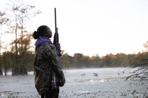 Young Woman In Hunting Gear Stands in wet-marsh at dusk.