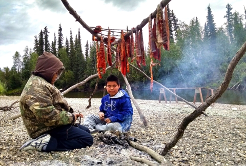 Two youngsters smoke salmon at a camp fire.