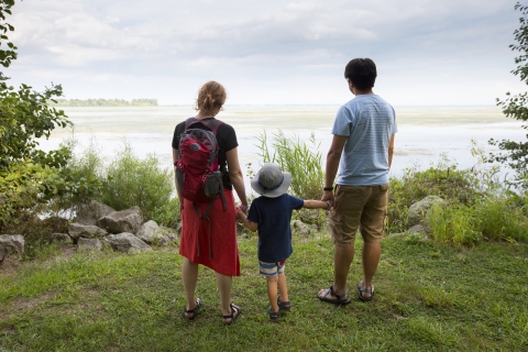 Two adults hold the hands of a child between them and look out at the river from the banks of the Detroit River International Wildlife Refuge.