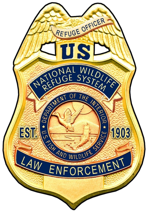 A light gold, shield-like badge with the words Refuge Officer, US, National Wildlife Refuge System, Department of the Interior, U.S. Fish and Wildlife Service, law enforcement, Est. 1903 on it in blue