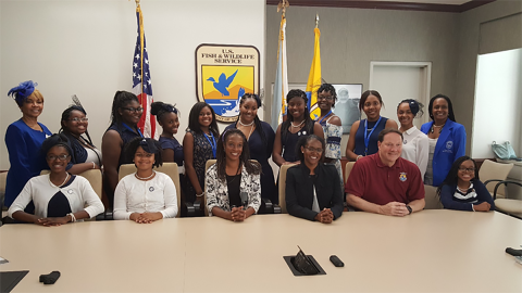 Group photo of Service employees and Zeta Phi Beta Sorority memebers sitting and standing at a long table with American flag and Service logo emblem in foreground. 
