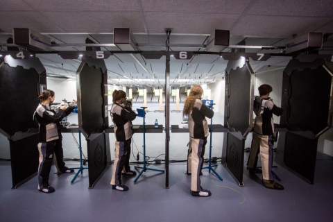 Four women standing from left to right aim their rifles at their targets at the end of the shooting range.