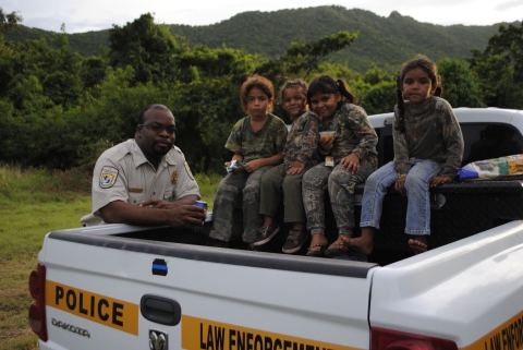 A uniformed Refuge System law enforcement officer leans against a flatbed police vehicle on which four smiling children sit.
