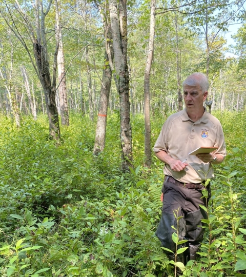 A man in wearing a USFWS uniform with a notebook in one hand and a pen in the other walks through a filed of wild plants towards the camera and away from a forest of tall tress.