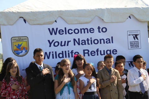 Kids in Sunday clothes with hands over heart in front of sign that says Welcome to Your National Wildlife Refuge