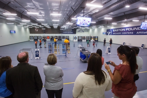 An audience standing behind a yellow line on the floor watch the archery competition from a safe distance .