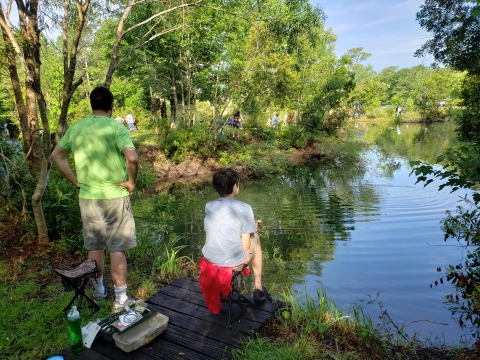 Father stands with hands over his waist while next to a sited kid holding a fishing pole. Both are facing a pond with their backs to the camera. Other people can be seen at a distance, fishing along the edge of the pond.