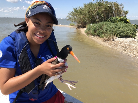 A smiling woman in a blue shirt holds a black and white bird with a long orange bill.