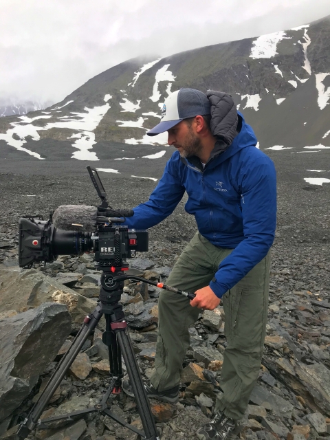 A man adjusting the angle of his large video camera while standing on a rocky glacial field with some snow on it