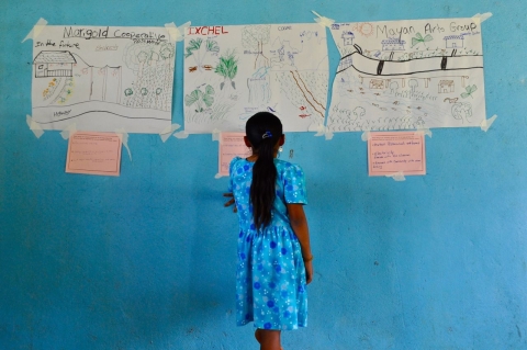A girl with black heair in a ponytail wearing a bright blue dress stands in front of a bright blue wall looking at hand-written village planning meeting documents taped to the wall