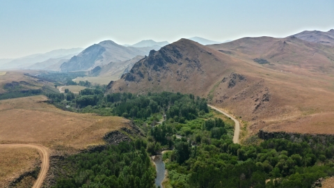 An aerial view of a stream surrounded by green vegetation and dry, brown mountains in the distance