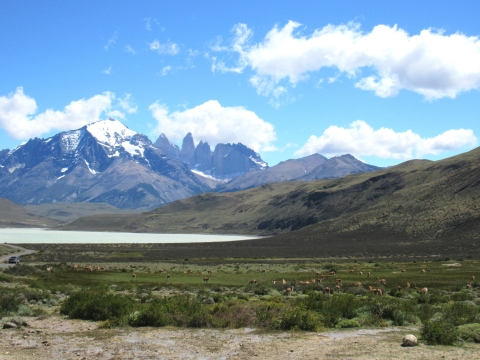 Mountain range with snow-capped peaks in background, and body of water, grass-covered hills, and field in foreground