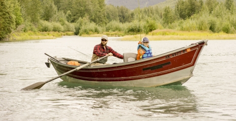A man paddles a boat with fishing rods along a river. The boat has another passenger.