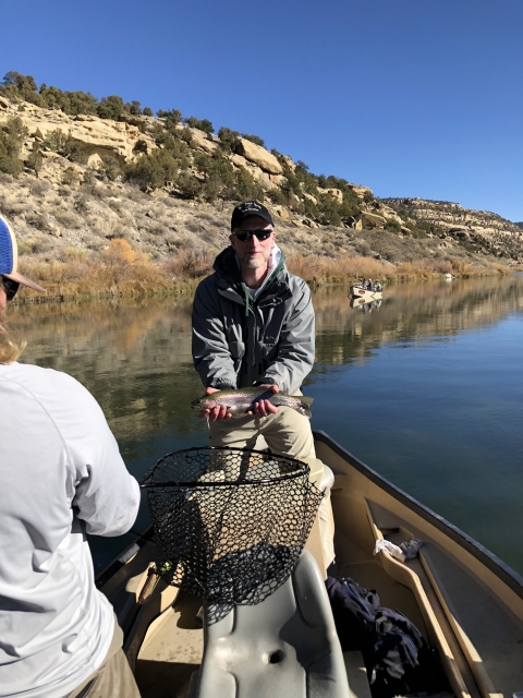 Man in boat on the water holding a fish. Man wears a black cap and sunglasses