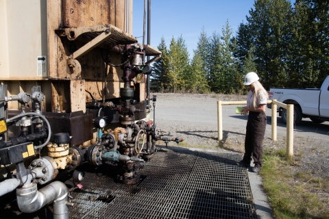 A woman in a hardhat and wearing a Fish and Wildlife Service uniform stands with a clipboard near a large piece of industrial equipment, which includes metal pipes