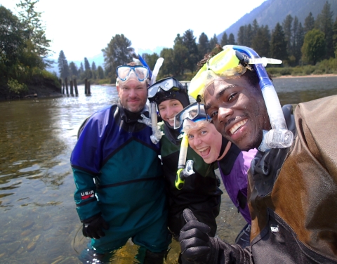 A group of 4 people in drysuits and snorkel masks stand in a river and grin for a selfie.