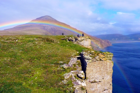 A rainbow span a grassy field, a steep cliff and blue water with a mountain in the background