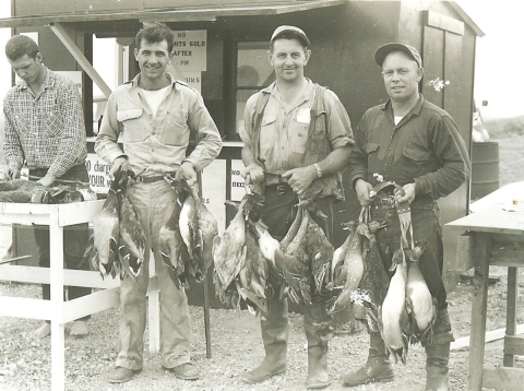 three hunters holding ducks. one check station staff member in background checking harvest. check station building in background