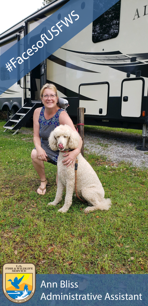 A woman crouches down in front of an RV next to a white poodle. A small grey bird is perched on her shoulder.