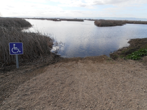 photo shows boat launch for mobility impaired hunters. blue and white mobility impaired sign on the left. gravel in foreground, water and vegetation in background