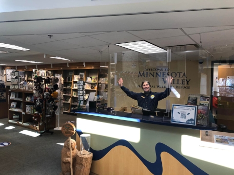 Volunteer Doris Ikier smiling and raising her arms wide while behind the information desk at Minnesota Valley National Wildlife Refuge's Bloomington Education and Visitor Center.
