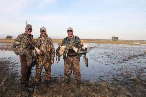 3 hunters holding harvested ducks and geese. water behind them
