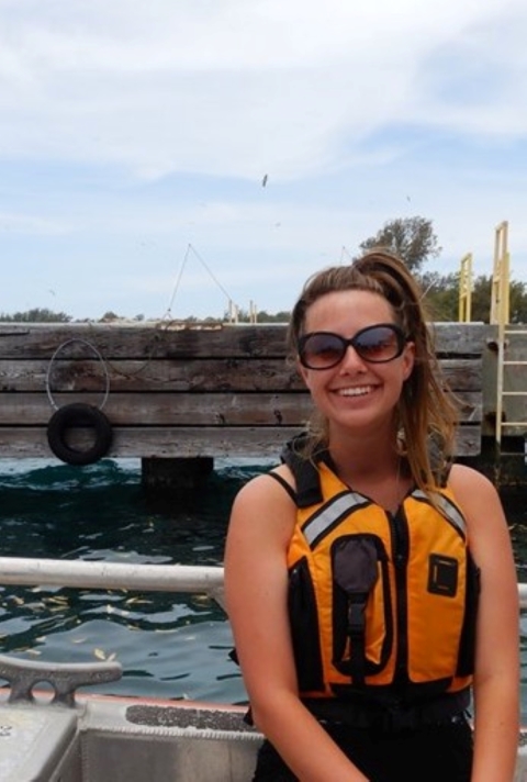 Gina McGuire looks smiles at the camera. She is wearing sunglasses and an orange lifejacket as she sits on a boat near a pier.