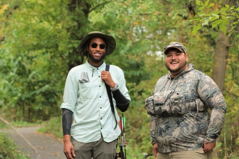 two people, one wearing a button down shirt, hat and glasses, and the other in camoflauge attire, pose in a forest smiling