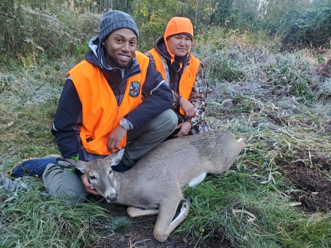 Two people in hunting jackets crouch next to a fallen deer