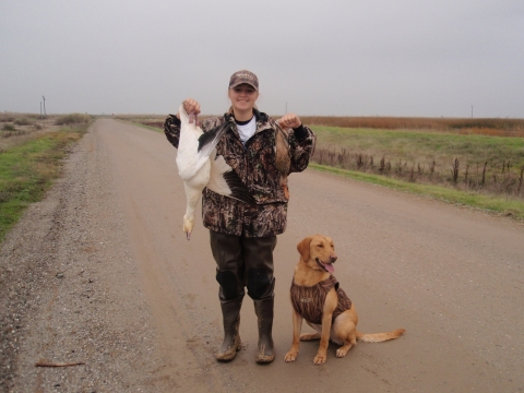 junior hunter holding up snow goose in one hand and duck in other hand. golden retriever dog sitting on person's left side