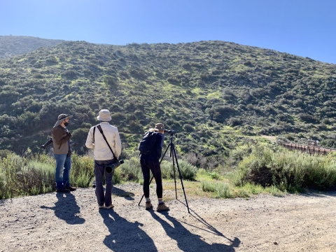 Three individuals standing on the trail looking towards a chaparral mountain. One person standing on the far right looks into a viewing scope. 