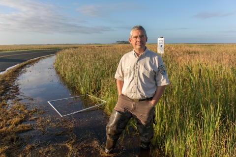 a man in Service uniform stands near a salt marsh with a refuge boundary sign behind him